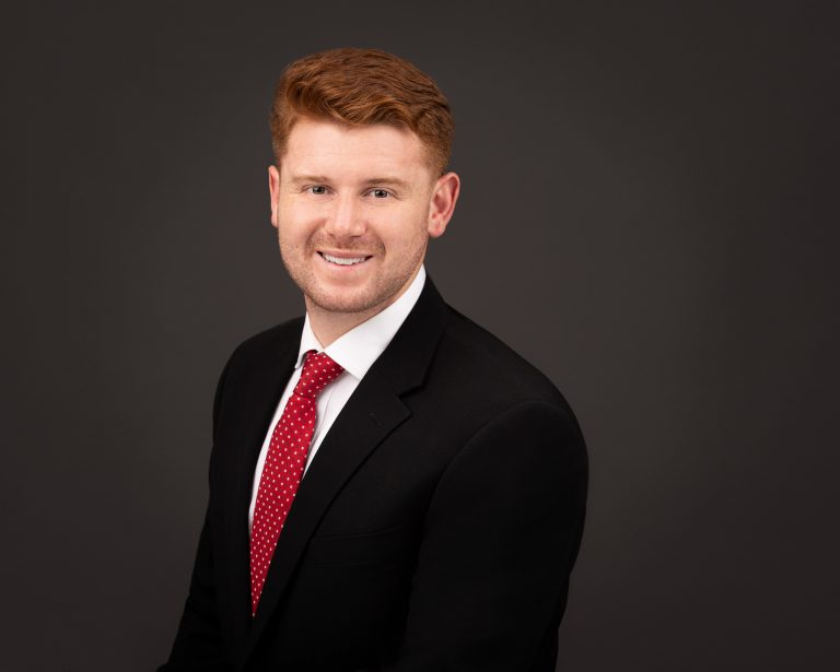 Executive Team Headshot on gray background of guy with red hair wearing black jacket and red tie